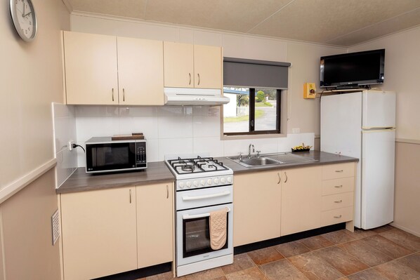 Cocina funcional con microondas, estufa, horno y refrigerador. Se observa una ventana con vista al exterior, un reloj en la pared y un televisor de pantalla plana sobre el refrigerador.