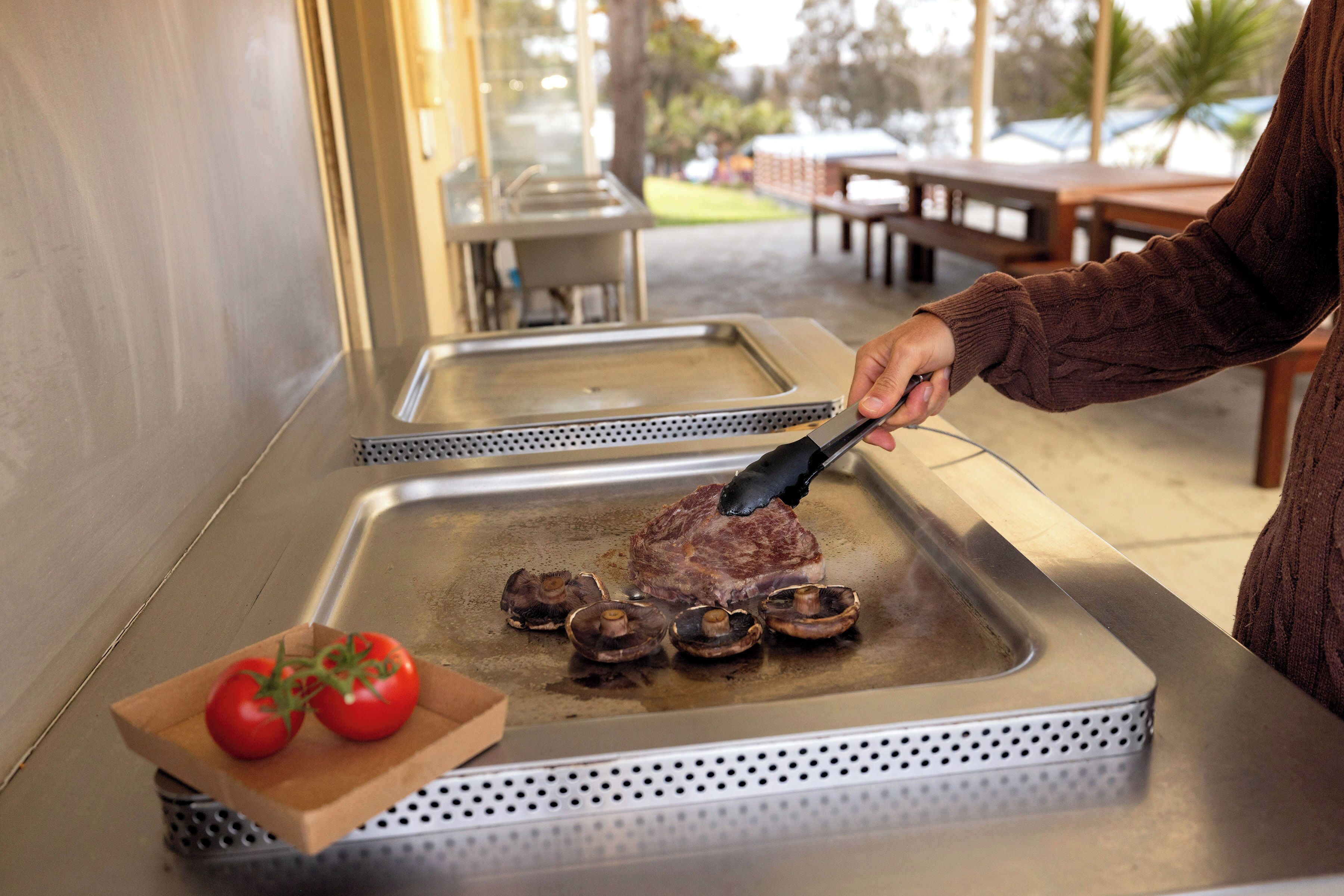 A person cooking a steak on a grill with mushrooms and tomatoes nearby.