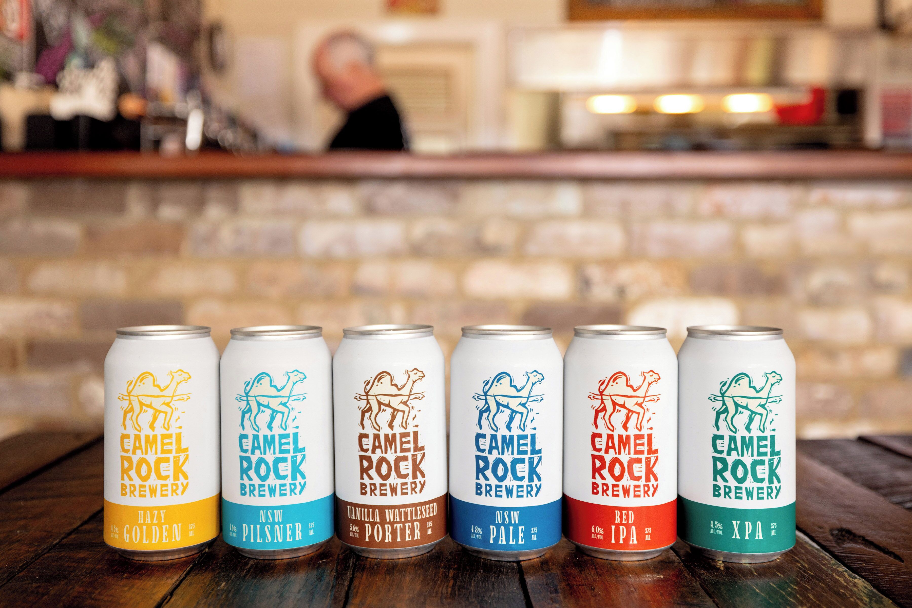 Seis latas de cerveza de Camel Rock Brewery sobre una superficie de madera.