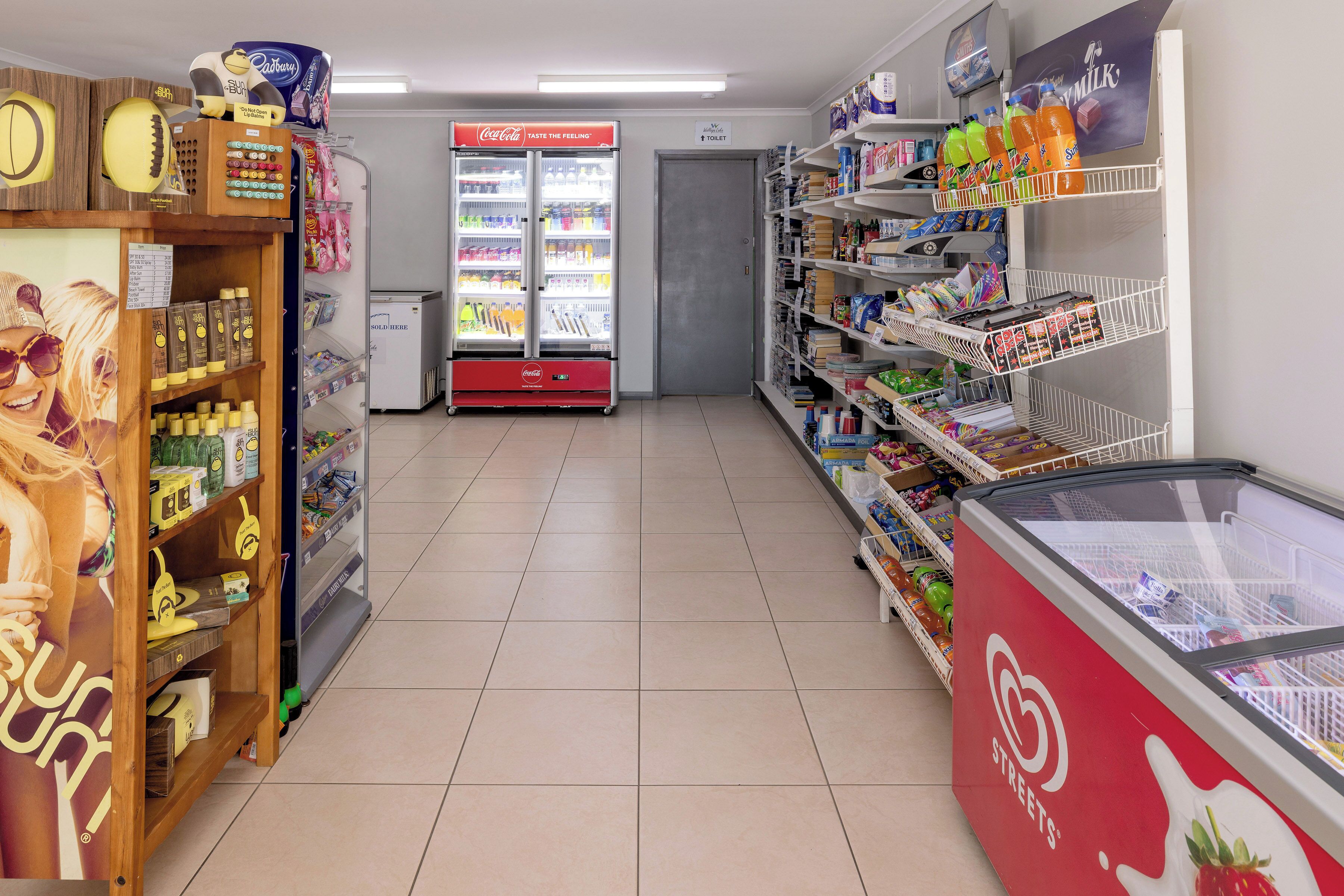 A grocery store aisle with shelves stocked with various products, a Coca-Cola refrigerator, and a freezer with a Streets logo.