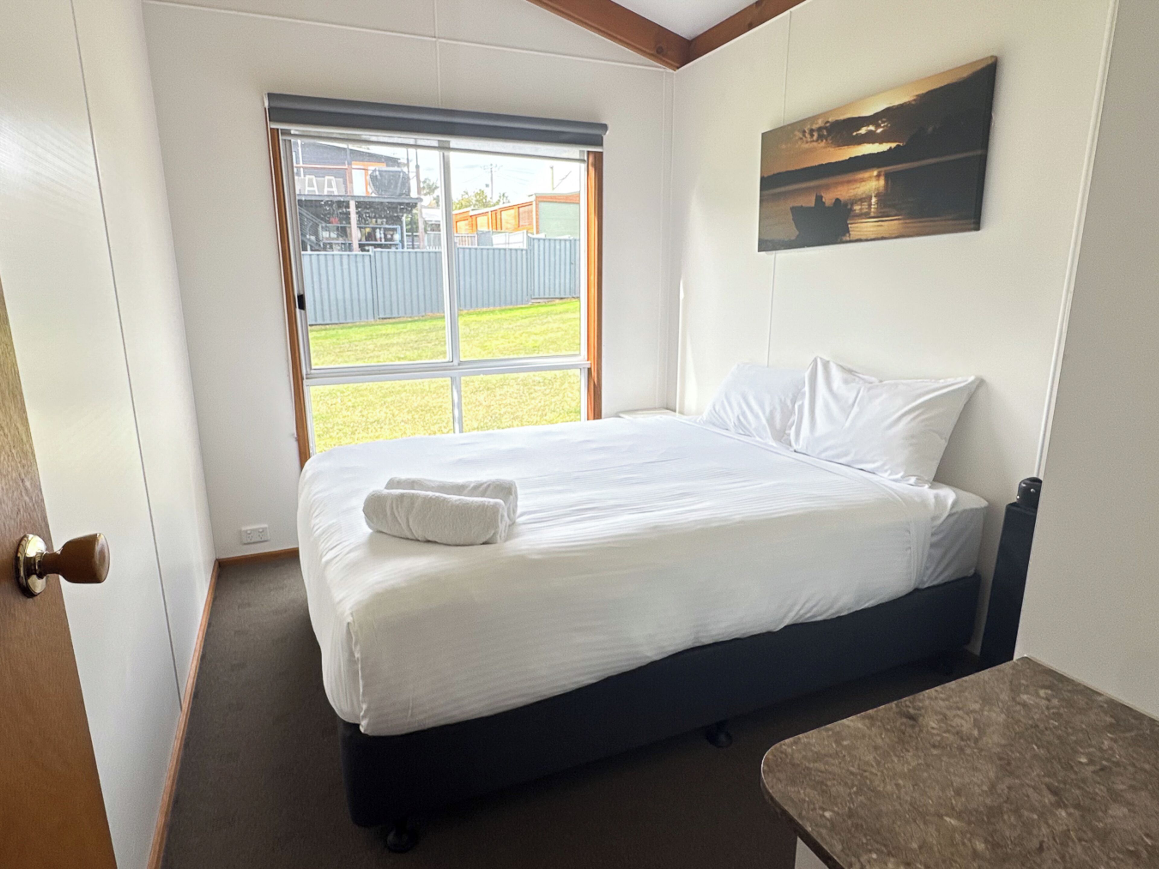 A hotel room with a bed, a window with a view of a building and a grassy area, and a painting on the wall.