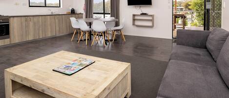 A modern living room with a grey sofa, a wooden coffee table, and a dining area with white chairs.