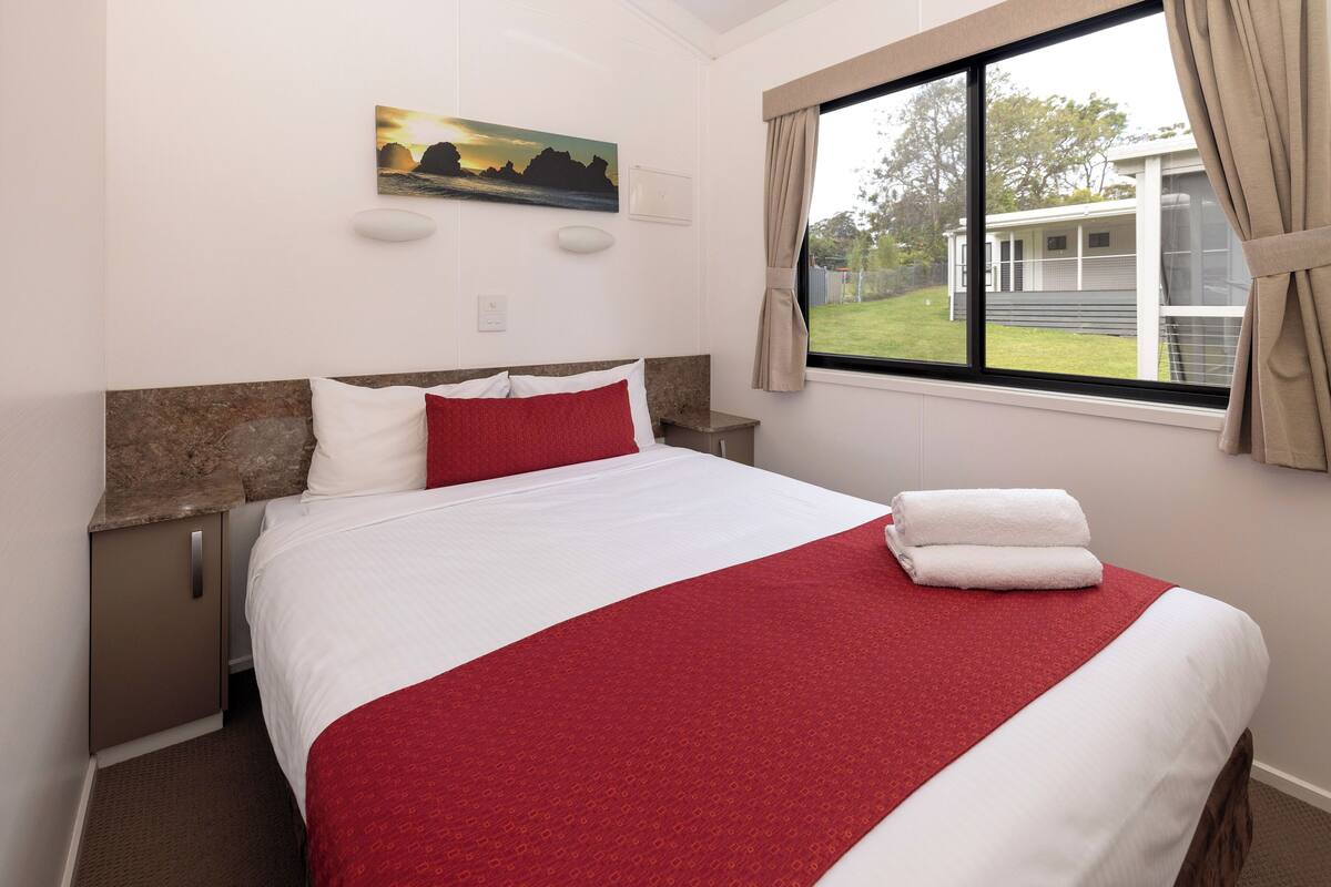 Een netjes opgemaakt bed met een rood-witte sprei, een schilderij aan de muur en een raam met gordijnen.