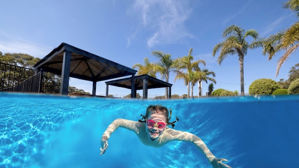 Une vue sous-marine d’une piscine à l’eau d’un bleu cristallin, avec un pavillon abrité et des palmiers en arrière-plan.