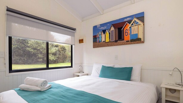 Une chambre d’hôtel avec un grand lit, une fenêtre donnant sur un paysage verdoyant, un tableau représentant des cabanes de plage et une table de chevet avec une lampe.