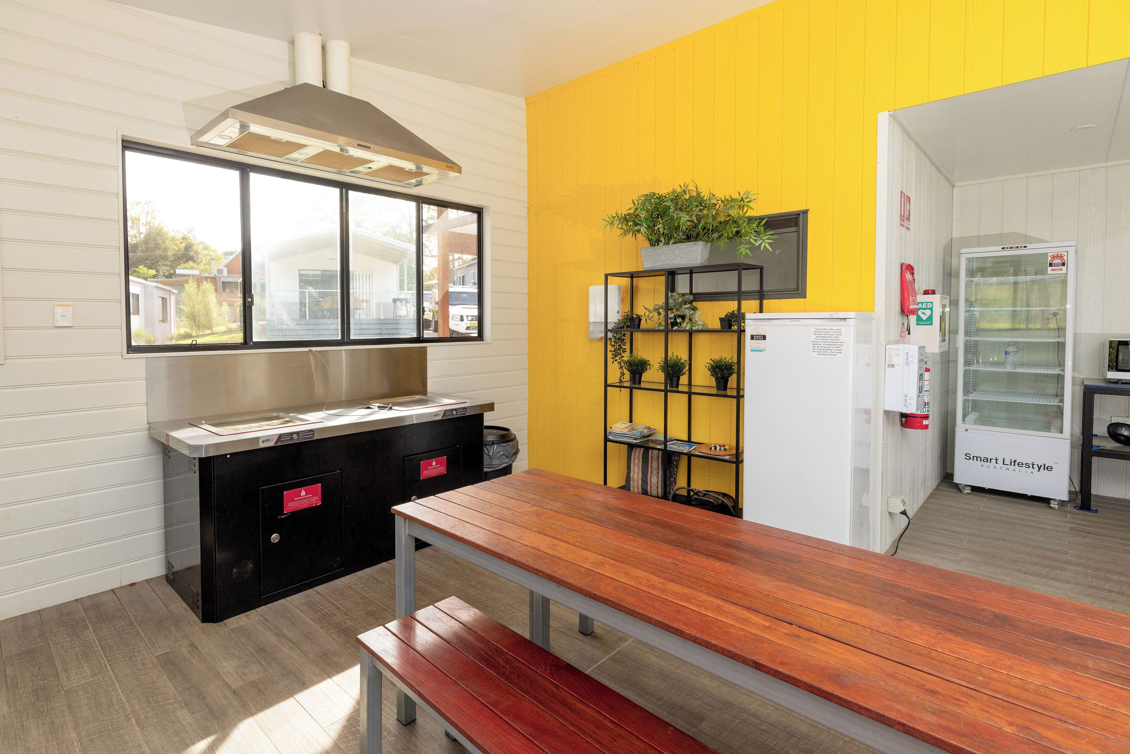 A kitchen with a wooden table and bench, a black countertop, a window with a view of houses, a yellow wall, and a white refrigerator.