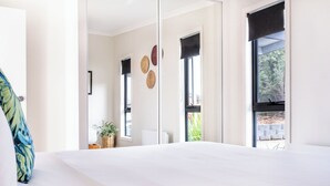 Un dormitorio con una cama grande, dos ventanas con persianas negras, una cesta de mimbre con una planta y un espejo en la pared.