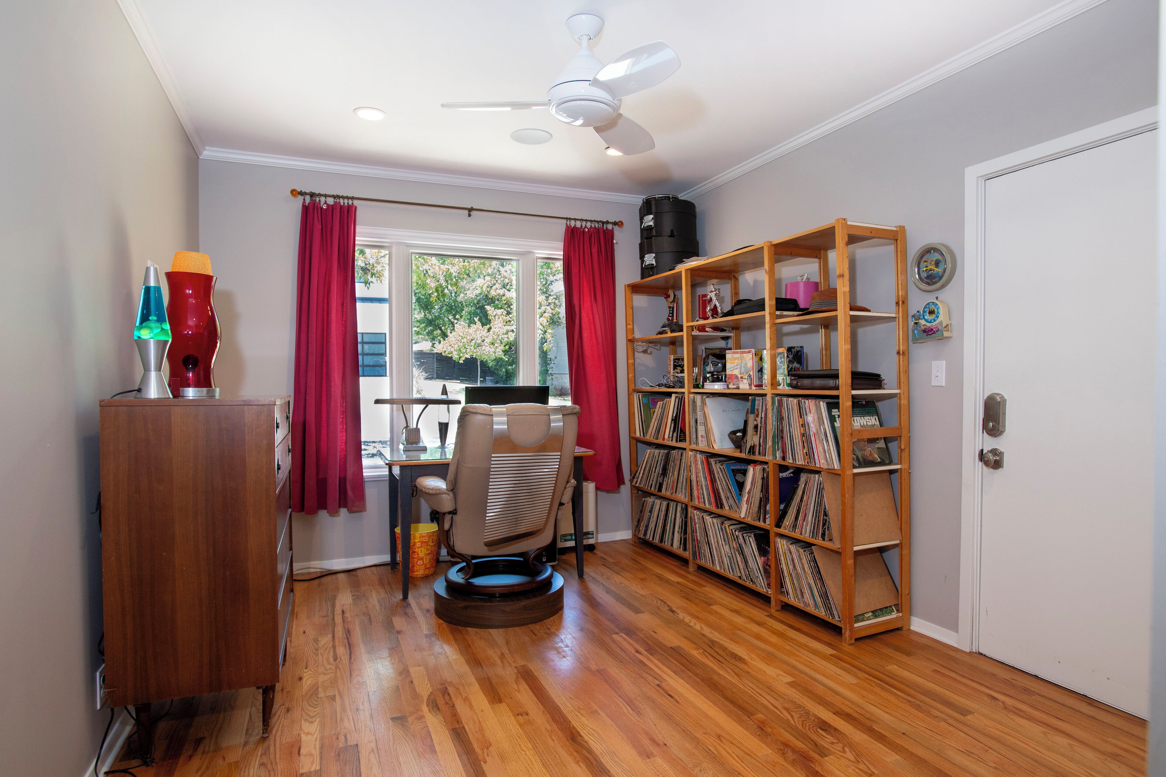 Ein Zimmer mit Holzboden, einem Schreibtisch mit Stuhl, einem Bücherregal und einem Fenster mit roten Vorhängen.