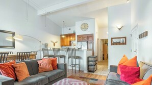 Ein modernes Wohnzimmer mit einem grauen Sofa, orangefarbenen Kissen und einem gemusterten Hocker. Im Hintergrund ist eine Küche mit Holzschränken und einem Kühlschrank zu sehen.