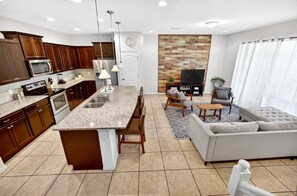 A modern kitchen with granite countertops, wooden cabinets, and stainless steel appliances. There is a dining area with a gray sofa and a coffee table.