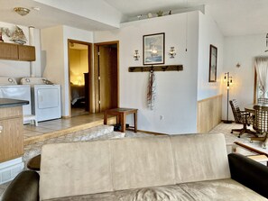 A living room with a beige sofa, a wooden coffee table, and a dining area with a checkered tablecloth and chairs. There is a laundry area with washing machines and a small stool.