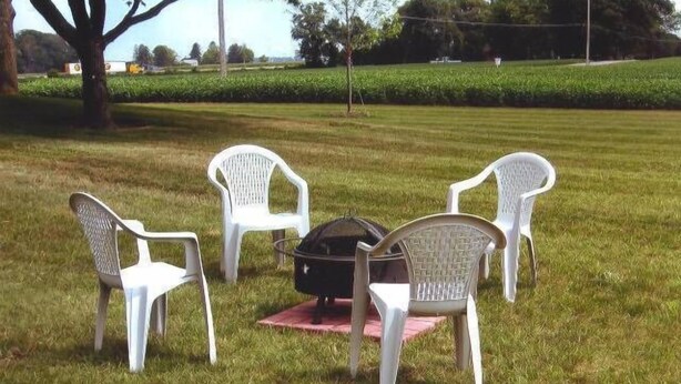 Quatre chaises en plastique blanc sont disposées autour d’un barbecue noir, sur un terrain herbeux.