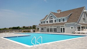 A swimming pool with a tiled deck, a covered seating area, and a large house with a gabled roof in the background. -  Serene Sanderling Beach Cottage Steps from beach w/community pool - Duck  (Duck)