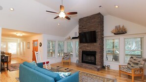 A spacious living room with a stone fireplace, a ceiling fan, and large windows with blinds. -  Serene Sanderling Beach Cottage Steps from beach w/community pool - Duck  (Duck)