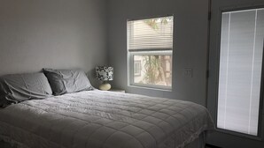 A bedroom with a bed, a nightstand, a lamp, a window, and a door. - Fay Cottage - Convenience in the heart of La Jolla village of La Jolla (La Jolla)