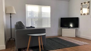A modern living room with a grey sofa, a small round table, a floor lamp, a TV on a white cabinet, and a large window with blinds. - Fay Cottage - Convenience in the heart of La Jolla village of La Jolla (La Jolla)