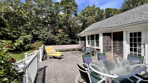 A covered outdoor deck with a glass table and chairs, a bench, and a view of a wooded area.