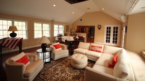 A spacious living room with a high ceiling, large windows, and a kitchen in the background.