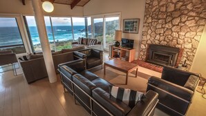 A spacious living room with a stone fireplace, leather sofas, a television, and a large window overlooking the ocean. - King George Beach Retreat (Middle River)