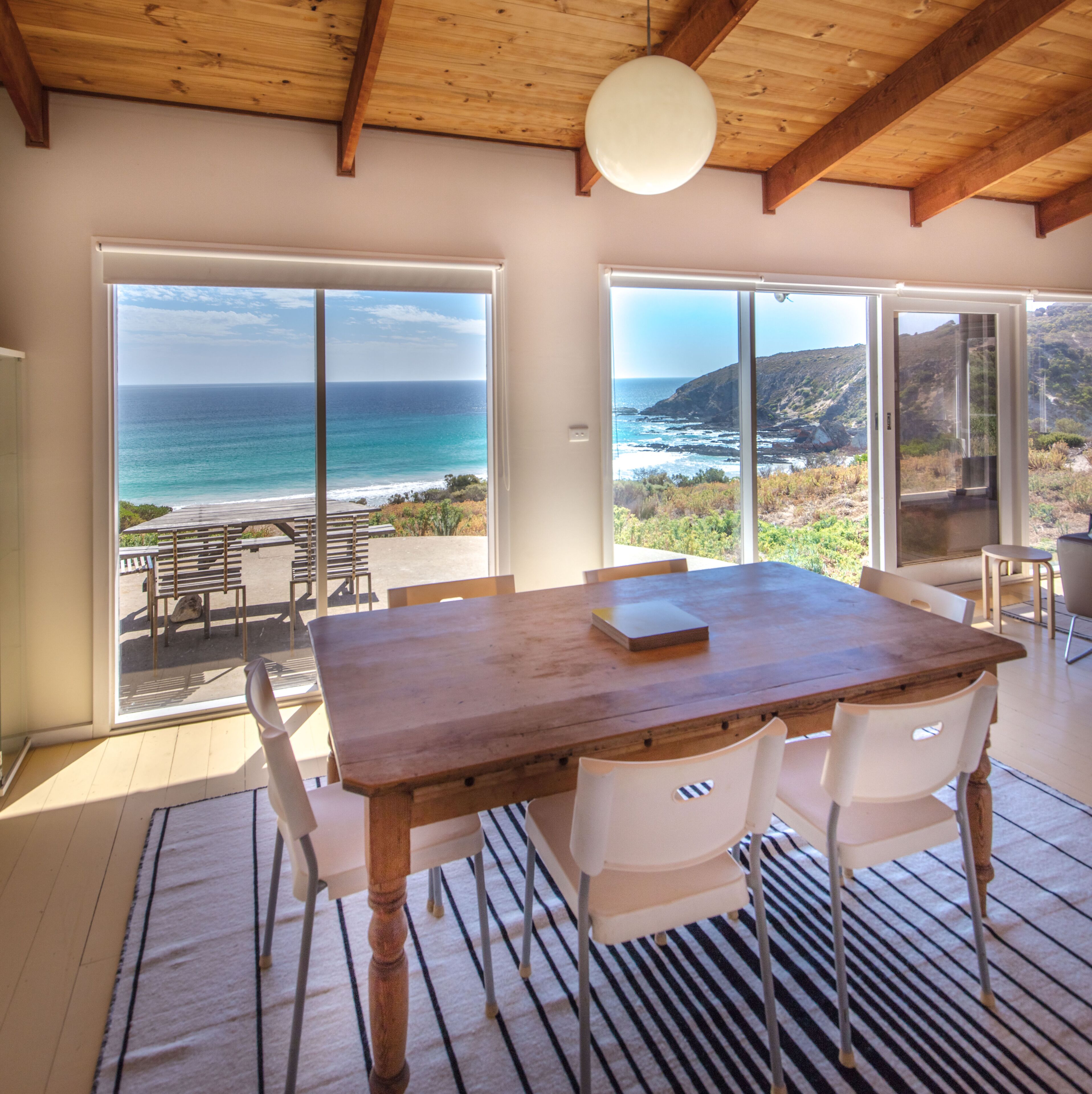 A wooden dining table with white chairs, large sliding glass doors leading to a patio with a view of the ocean and hills.