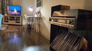 A living room with a flat-screen TV, a table with a lamp, a record player, and a bookshelf. - Downtown Loft: King/Queen Suites- 2½ Baths  Near Convention Center EV charging (Fort Smith)