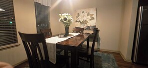 A dining area with a wooden table, chairs, a vase with flowers, and a painting on the wall.