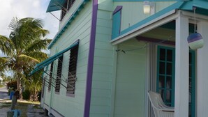 Una casa de dos pisos con un porche y una pequeña terraza, ubicada en una zona arenosa con palmeras al fondo.