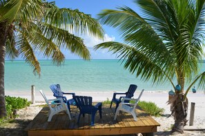 Un pontile di legno con due sedie blu e una bianca, affacciato su una spiaggia con acque cristalline turchesi e palme.