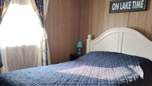 Ein Schlafzimmer mit Bett, Nachttisch, Lampe, einem Fenster mit Vorhängen und einem Wandschild mit der Aufschrift 'ON LAKE TIME'.