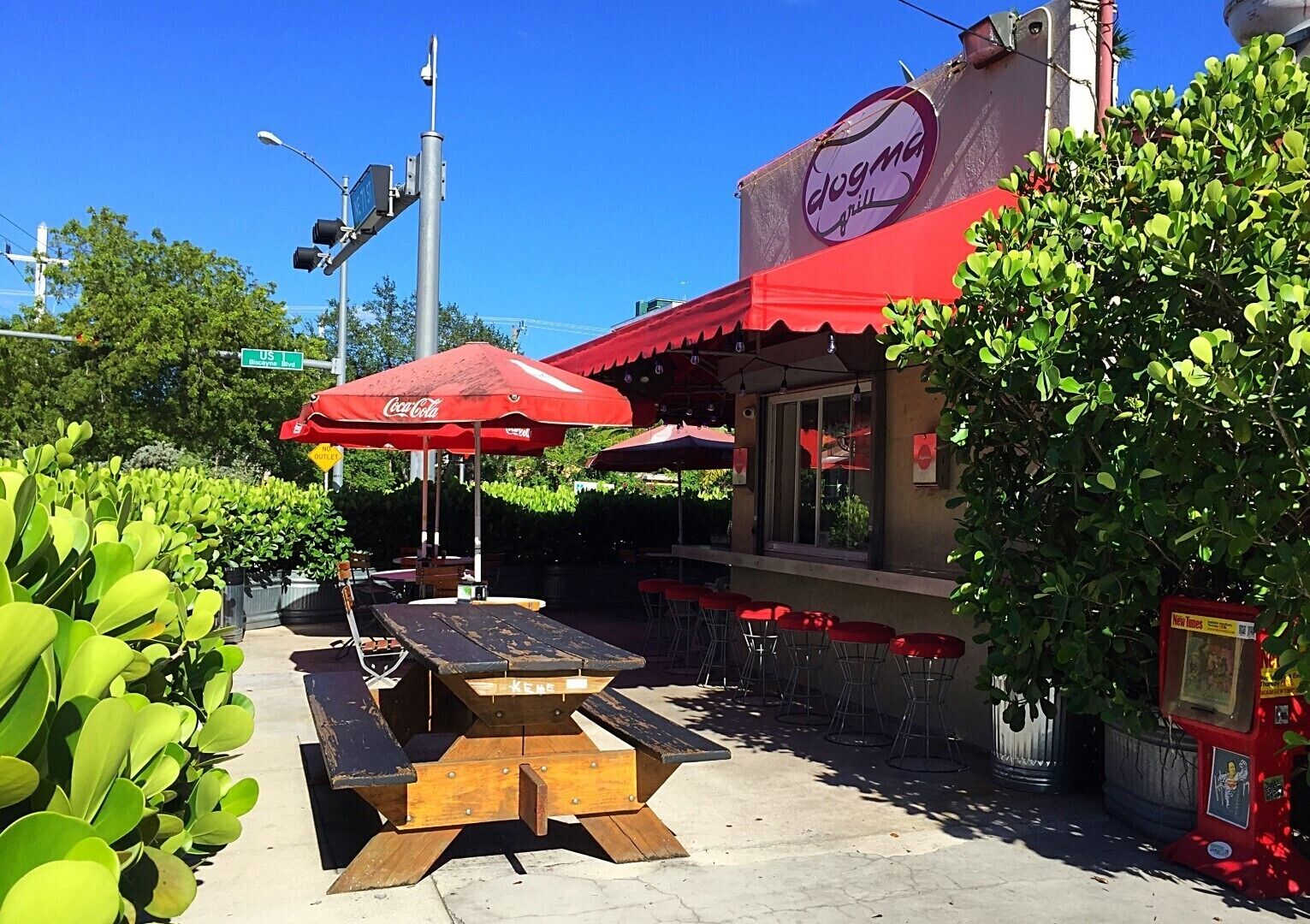 An outdoor seating area with tables and red umbrellas, a Coca-Cola sign, and a 'Dogma Grill' sign.