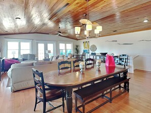 A spacious living room with a wooden ceiling, a dining area with a wooden table and chairs, and a large window providing natural light.