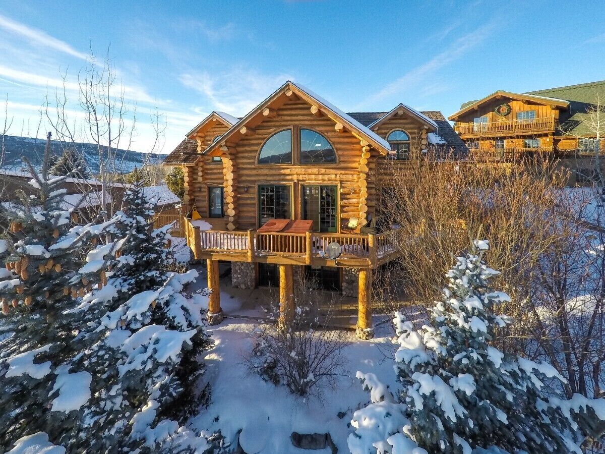 Une cabane en rondins dotée d’un balcon et d’une terrasse, entourée d’arbres couverts de neige.