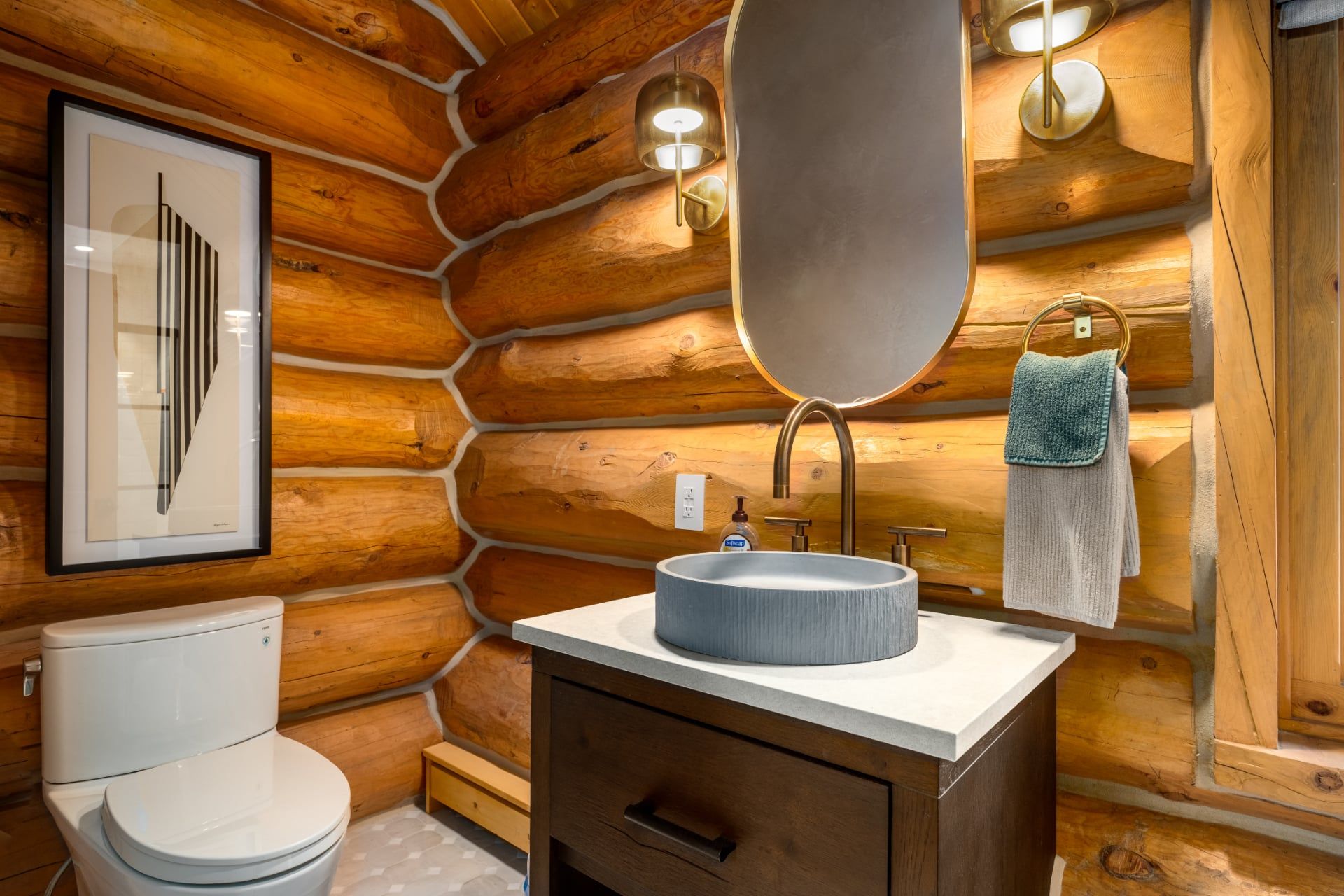 Ein Badezimmer mit einer Wand aus Holzstämmen, einer weißen Toilette, einem runden Waschbecken mit Spiegel und einem Handtuch an einem Handtuchhalter.