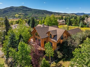 A large log cabin with a deck, surrounded by lush greenery and a mountainous backdrop.