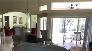 A spacious living area with a grey sofa, a dining table, and chairs. There is a large window providing natural light and a view of the outdoors.