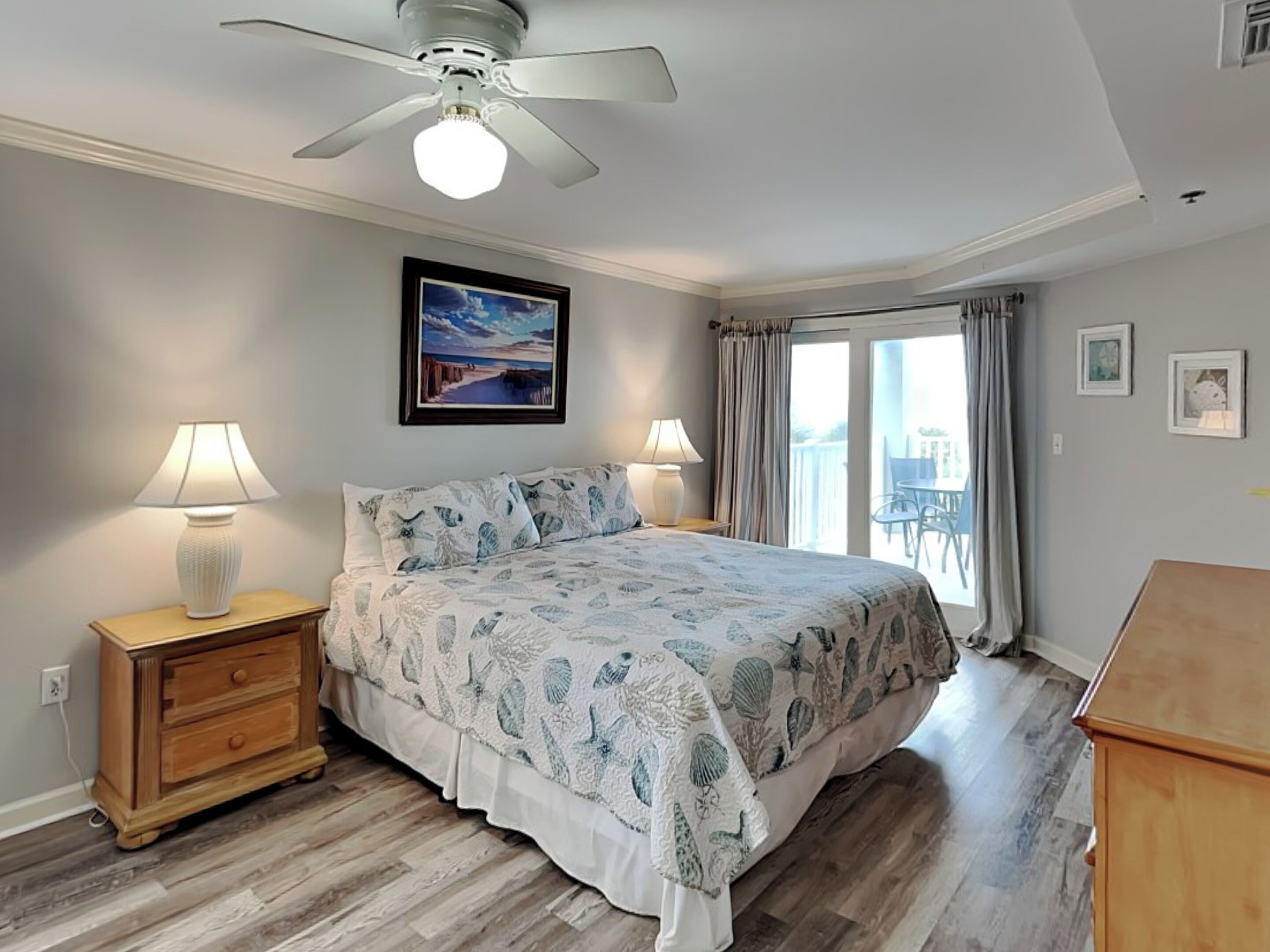Une chambre à coucher comprenant un lit, deux tables de chevet, un ventilateur de plafond, un tableau et une vue sur l’extérieur, accessible par une porte-fenêtre coulissante.