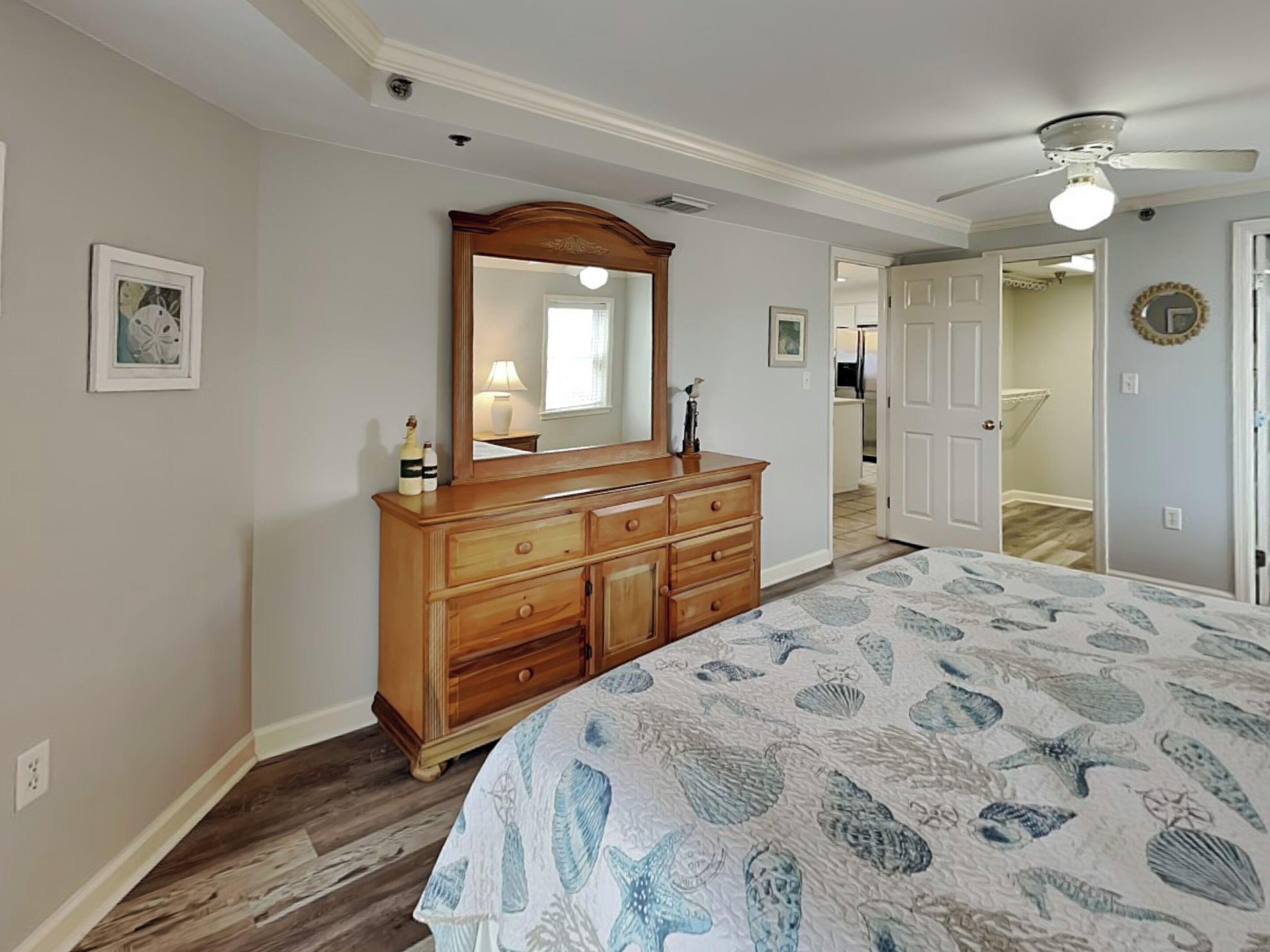 Un dormitorio con un aparador de madera, una cama grande con una colcha estampada con motivos de conchas marinas, un ventilador de techo y una puerta que da a otra habitación.