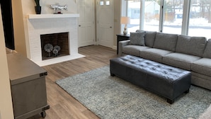Ein Wohnzimmer mit Kamin, einem grauen Sofa, einem grauen Hocker und einem kleinen Tisch mit einer Lampe.