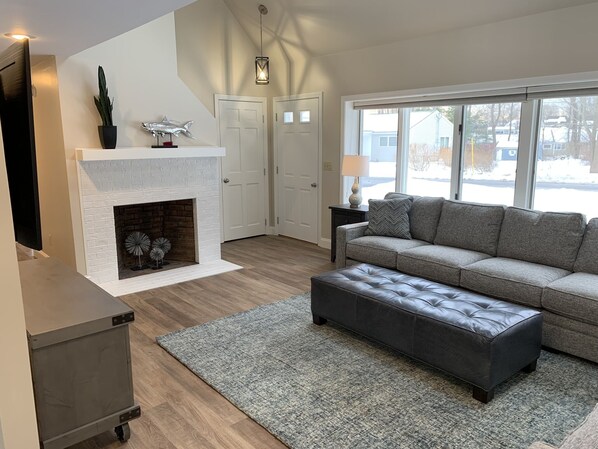 A living room with a fireplace, a grey sofa, a grey ottoman, and a small table with a lamp.