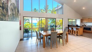 A spacious kitchen with large windows, a dining area with a wooden table and chairs, and a view of the outdoors. - Cooinda Gardens 1, Beautiful Apartment on Hamilton Island (Hamilton Island)