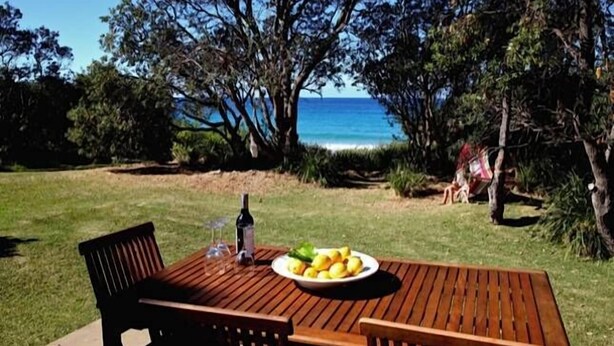 A wooden table with a bowl of lemons and a bottle of wine, set on a lawn with a view of the sea and trees. - ABSOLUTE Mollymook BEACHFRONT- Direct beach access  and views / Wifi / Smart TV  (Mollymook Beach)