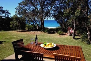Una mesa de madera con un bol de limones y una botella de vino, sobre un césped con vistas al mar y a los árboles.