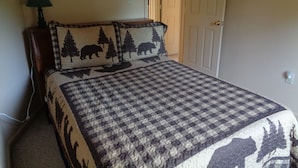 Quarto com colcha xadrez, duas almofadas com desenho de urso e árvore, abajur e porta à direita.
