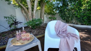 Een terras met een tafel gedekt voor drankjes, een witte stoel met een roze doek, en op de achtergrond bomen en een witte muur.