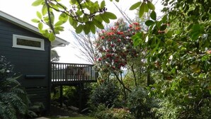 A residential garden with a wooden deck, lush greenery, and a house with a dark exterior. - Tui Glen - Lake front bach with stunning views (Lake Tarawera)