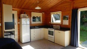 A compact kitchen with a microwave, stove, and sink, wooden walls, and a window with a view of greenery. - Tui Glen - Lake front bach with stunning views (Lake Tarawera)