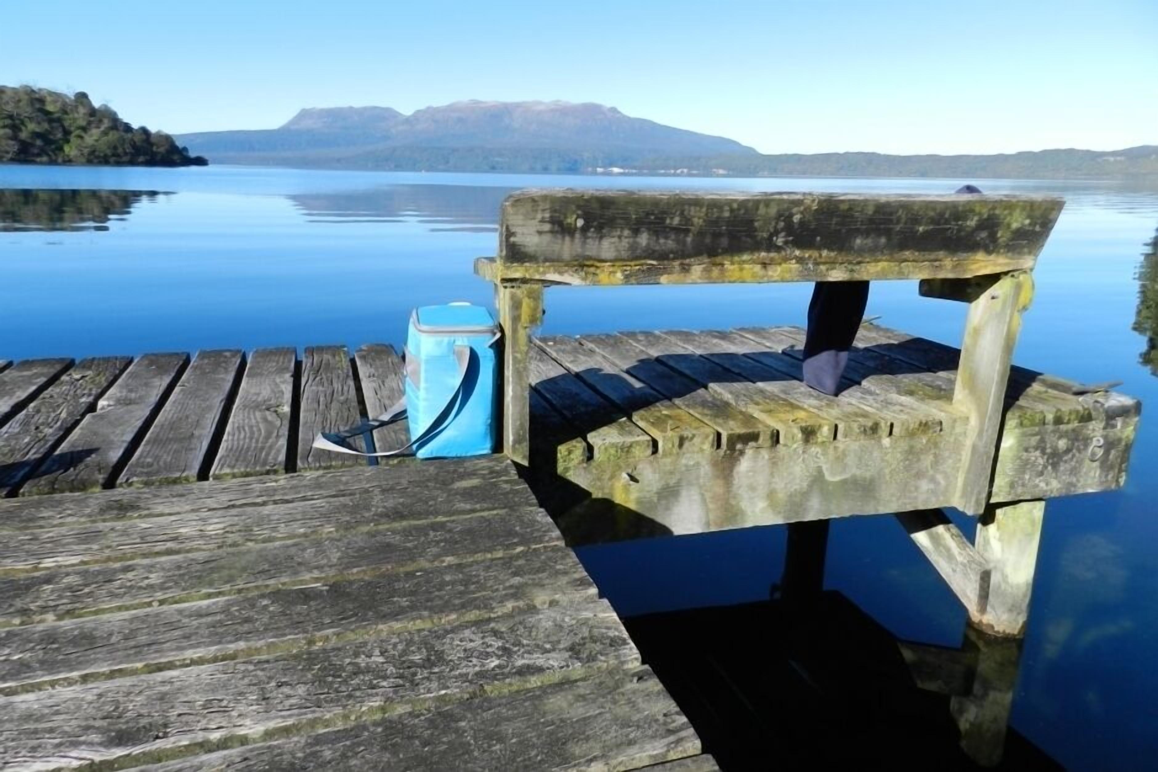 A wooden dock extending into calm water with a bench and a blue container.