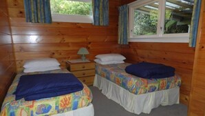 A wooden cabin bedroom with two beds, a nightstand, a window with curtains, and a view of greenery outside. - Tui Glen - Lake front bach with stunning views (Lake Tarawera)