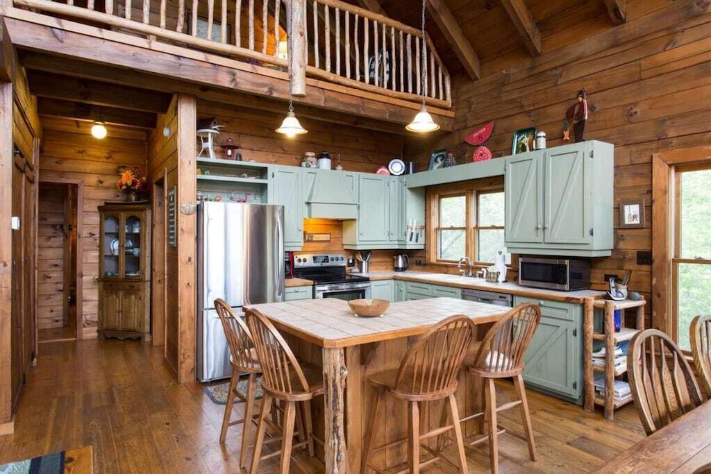 A rustic cabin with a wooden interior, a central island table, and a balcony.