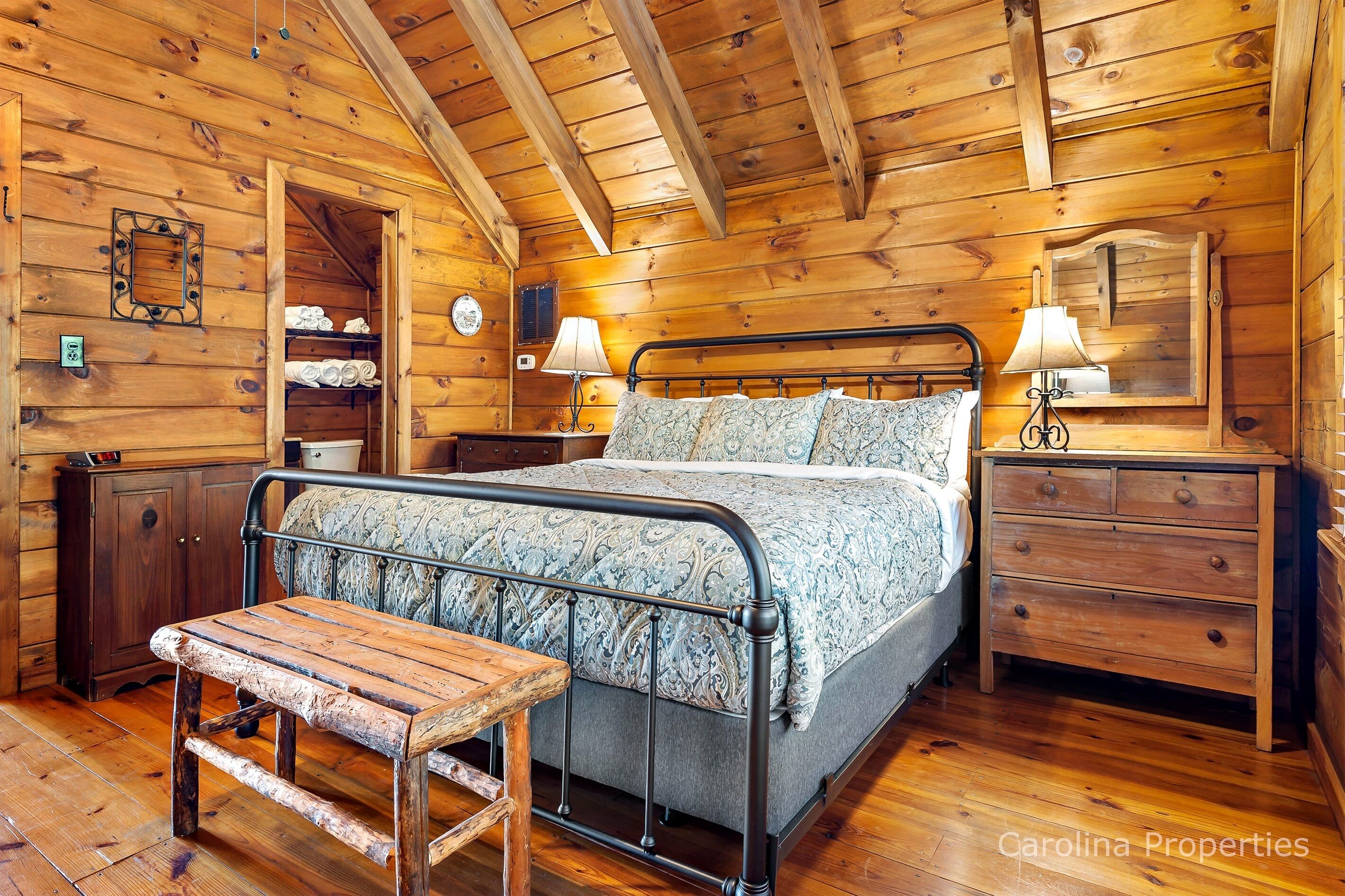 Une chambre dans une cabane en bois, avec un lit en métal, un banc en bois et une commode dotée d’un miroir.